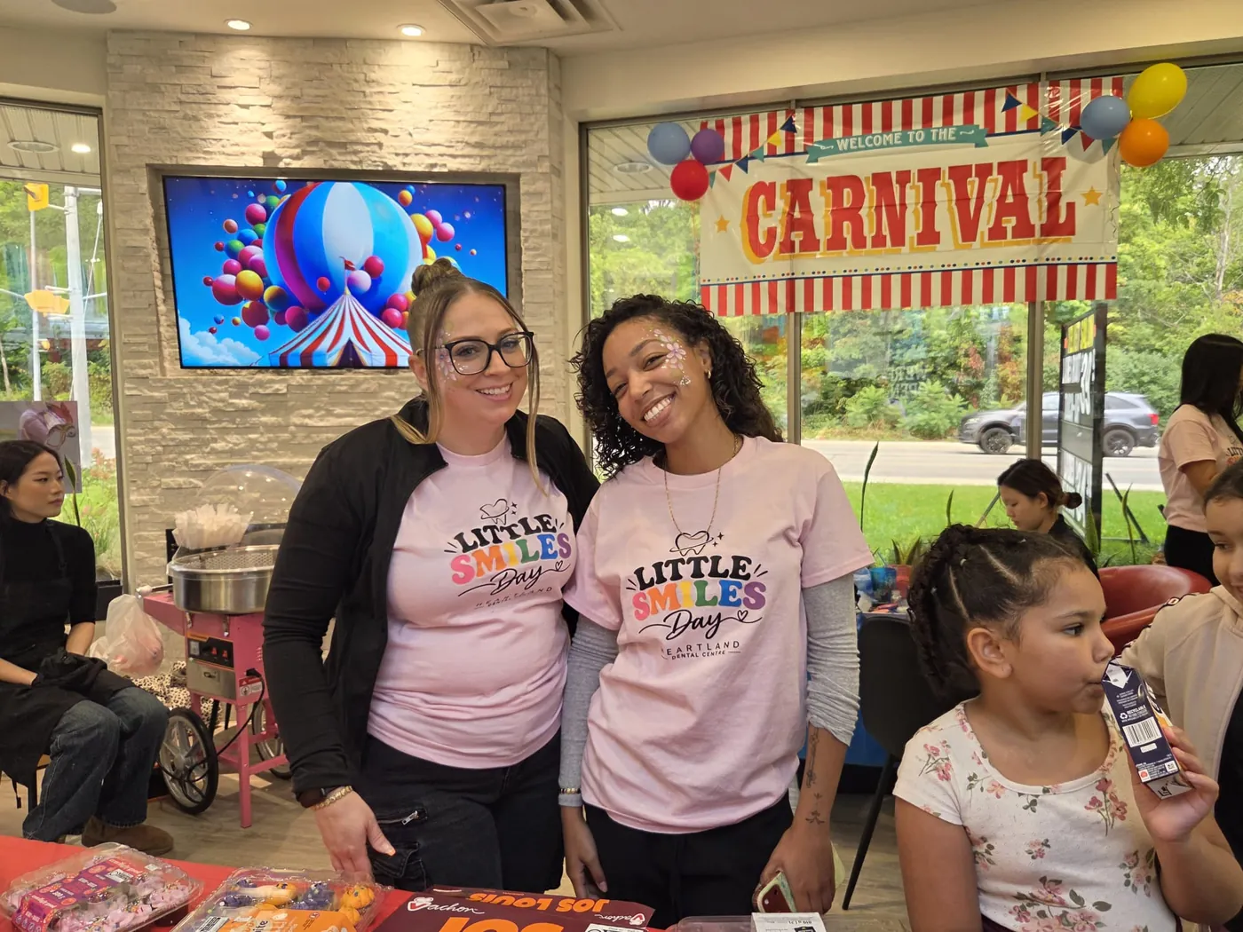 Two Heartland team members in Little Smiles Day shirts smiling in front of the carnival banner